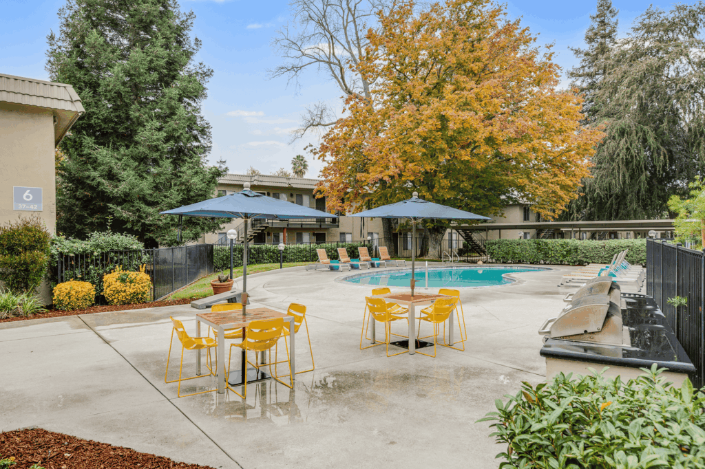 Outdoor pool area with yellow chairs and tables under blue umbrellas, surrounded by trees with autumn foliage and apartment buildings in the background. Poolside lounge chairs line the curved pool.