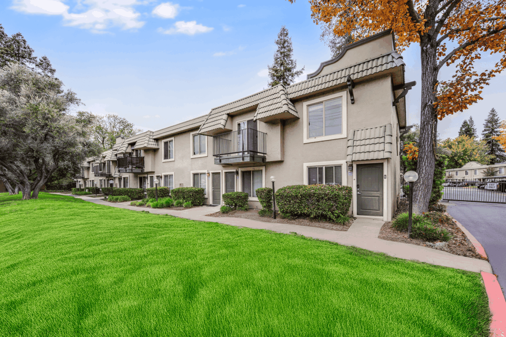 Two-story tan apartment buildings with balconies and patios face a well-manicured green lawn. Trees with autumn leaves and a pathway line the front, under a bright blue sky with scattered clouds.
