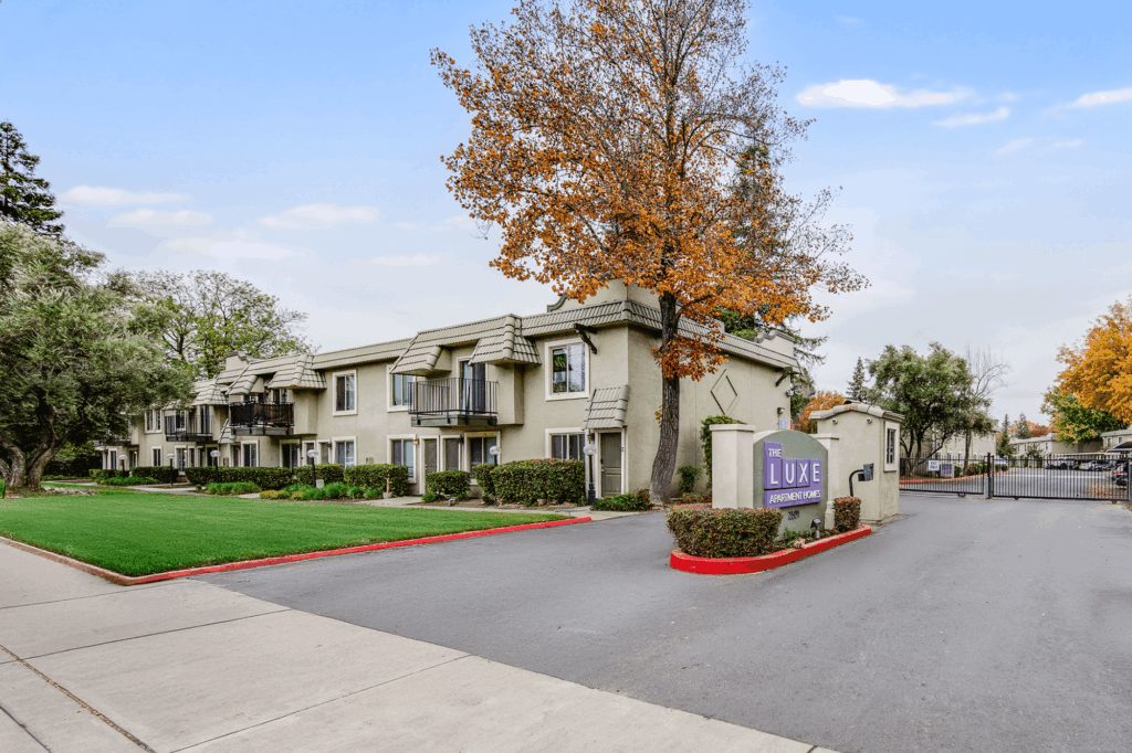 A two-story beige apartment building with balconies, surrounded by green lawns and trees with autumn leaves. A sign near the entrance reads Luxe Apartment Homes next to a gated driveway.