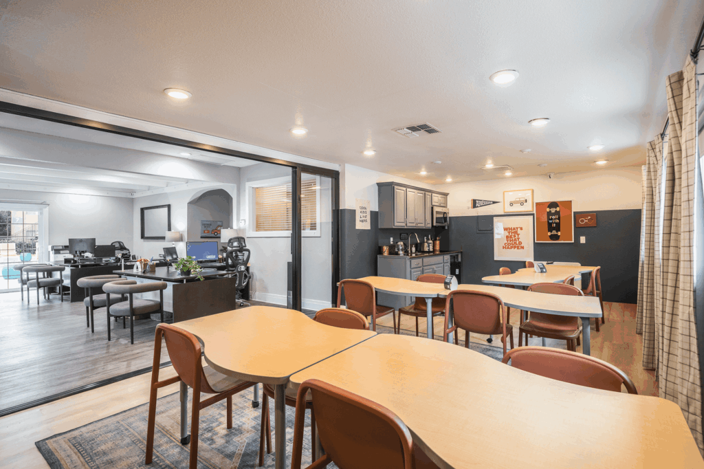 A spacious, modern room with tables and chairs arranged for group work, a lounge area with desks and office chairs, a coffee station, and motivational posters on the walls. Large windows let in natural light.