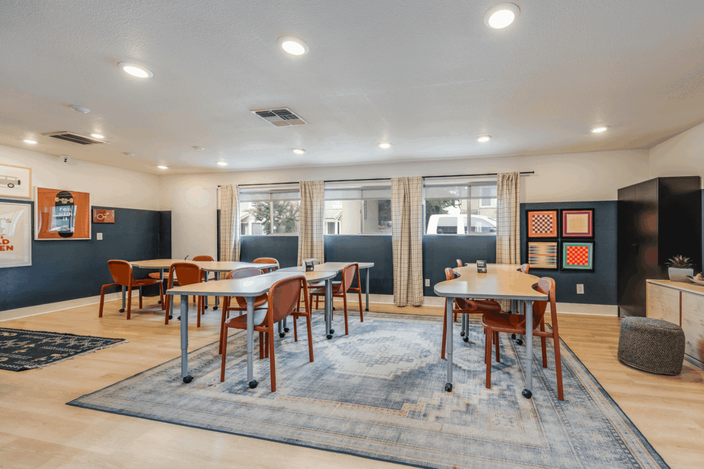 A classroom with several tables and orange chairs on a blue patterned rug, large windows with curtains, wall art, cabinets, and light wood flooring. The room is well-lit with recessed ceiling lights.