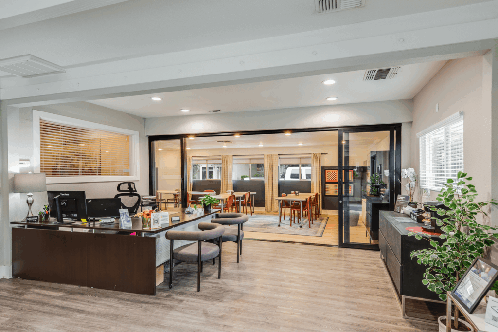 A modern office space with a desk, chairs, and office supplies in the foreground, separated by glass doors from a bright meeting room with tables, chairs, and large windows in the background.